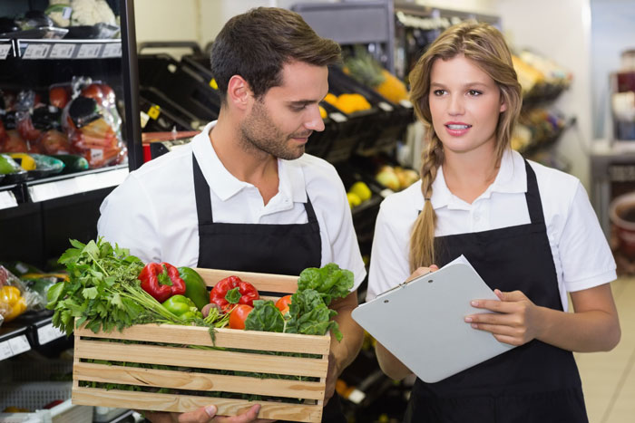 Two grocery store workers wearing aprons, one holding a crate of fresh vegetables, discussing tasks inside the store.