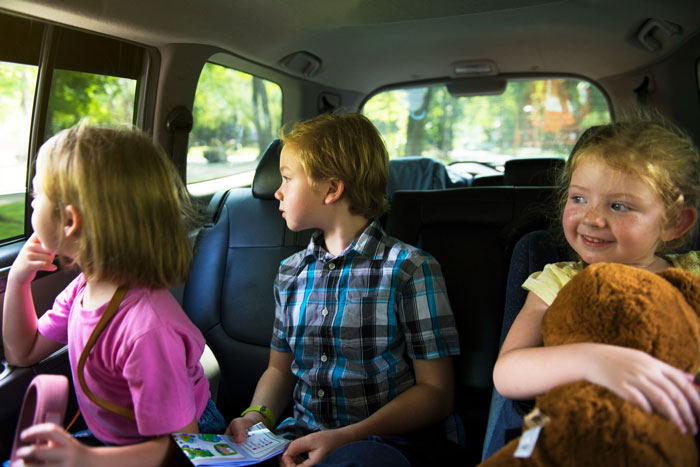 Three young children in a car, one holding a book and another smiling while hugging a teddy bear, highlighting guilt after calling CPS.