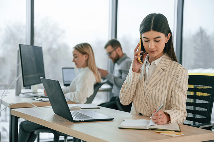 Young professional woman talking on phone and taking notes in office while coworkers work on computers nearby Young professional woman talking on phone and taking notes in office while coworkers work on computers nearby