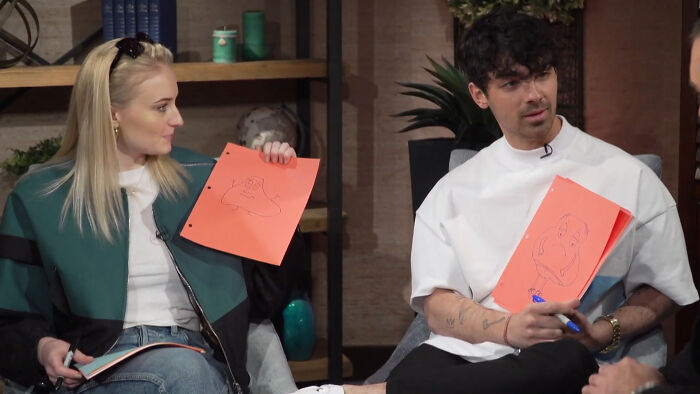 Two people casually sitting indoors, holding up orange papers with drawings, sharing a light moment about celebrities.