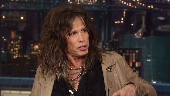 Rockstar with long curly hair and layered necklaces sharing stories about celebrity encounters on a late-night talk show.
