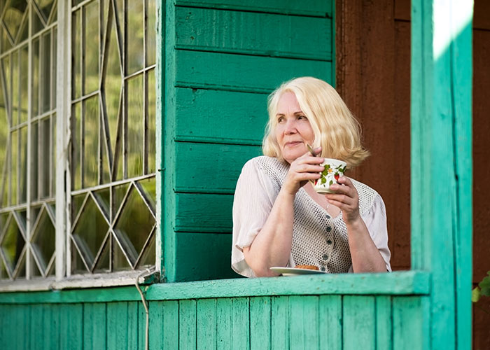 Older woman holding a cup on a porch, with a thoughtful expression, related to MIL poisoning family dog controversy.