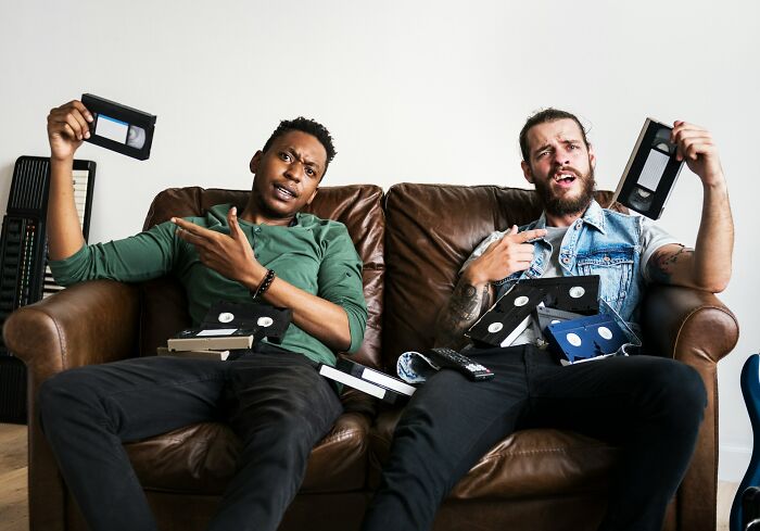 Two frustrated men sitting on a couch surrounded by video tapes, expressing moments that highlight I Don’t Get Paid Enough.