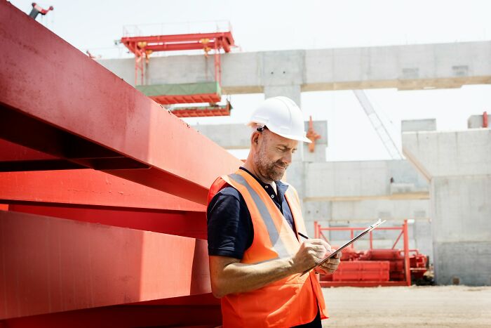 Construction worker wearing a hard hat and safety vest, taking notes at a building site, showcasing incredible minds without degrees.