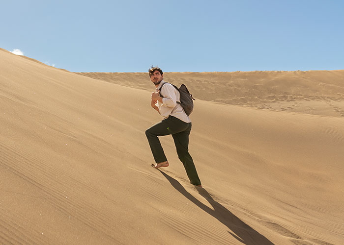 Man with backpack climbing steep sand dune in bright sunlight, illustrating survival hacks risks in desert environment.