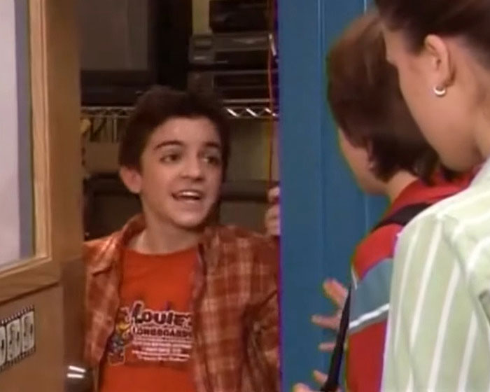 Nickelodeon star in casual clothes speaking to two others inside a school hallway scene from a TV show.