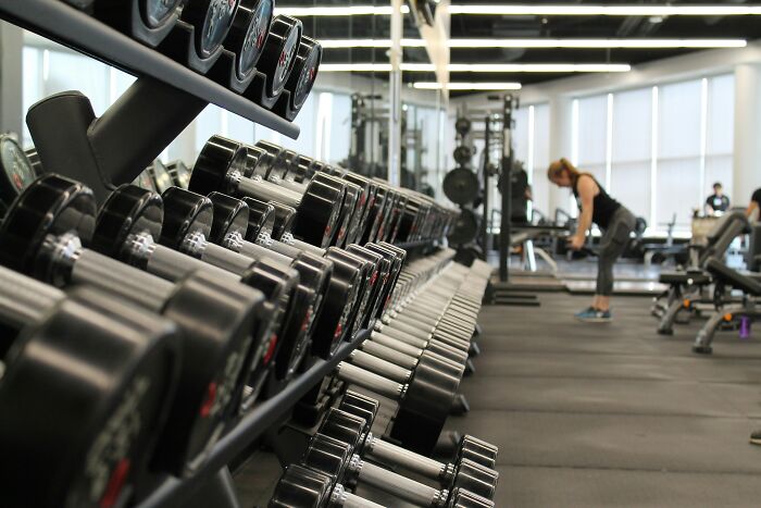 Rows of dumbbells in a gym with a woman exercising in the background promoting habits affecting mental and physical health