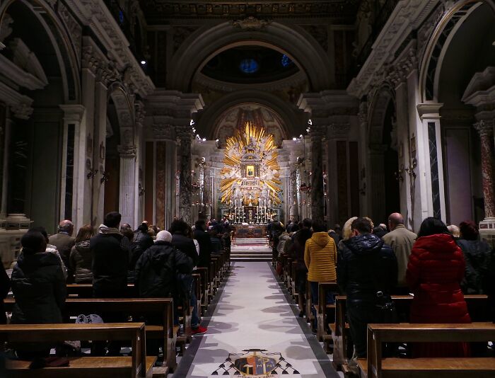 People attending a ceremony inside a grand church with ornate architecture and an illuminated altar in the background.