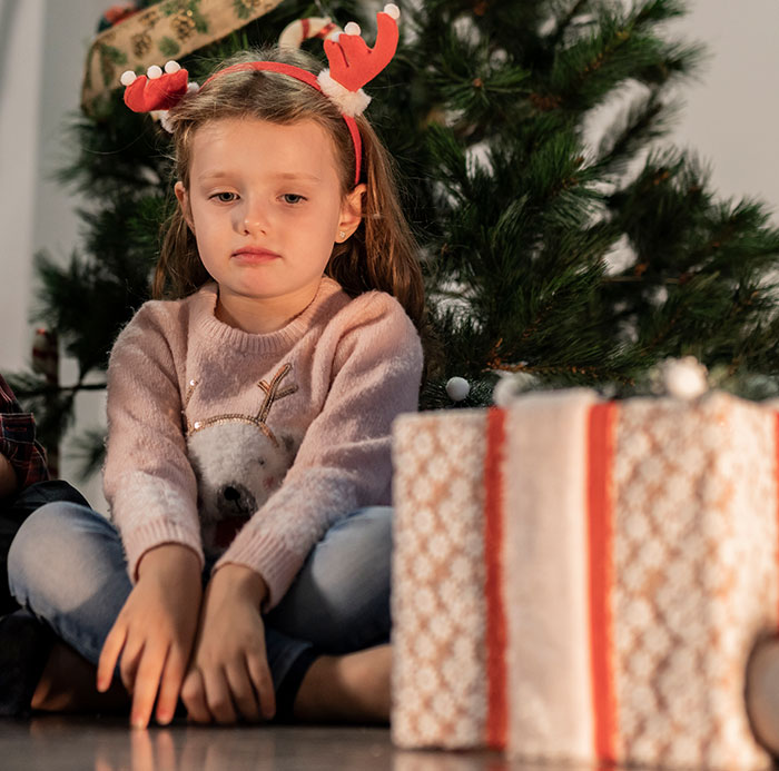 Sad 7-year-old girl with reindeer antlers sitting by Christmas tree and unopened gifts, reflecting holiday punishment debate. Sad 7-year-old girl with reindeer antlers sitting by Christmas tree and unopened gifts, reflecting holiday punishment debate.