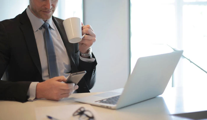 Man in suit drinking coffee while checking phone near laptop in bright office, related to gay daughter family conflict advice. Man in suit drinking coffee while checking phone near laptop in bright office, related to gay daughter family conflict advice.