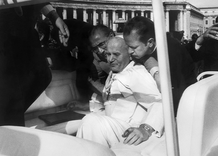 Black and white photo of Vatican officials assisting a man in white robes inside a vehicle, related to Vatican girl cold case.