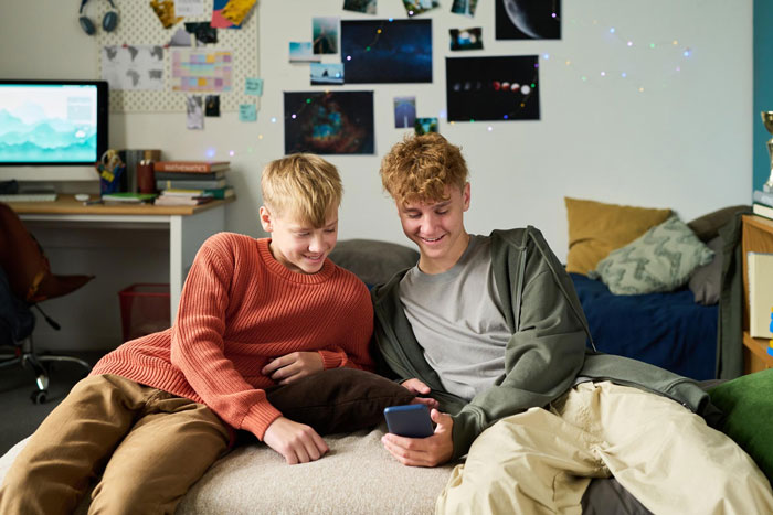 Two teenage boys sitting on a bed smiling at a smartphone, depicting teen mad as dad gifts stepbro a car for Xmas.