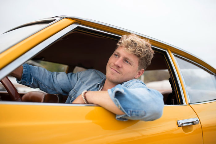 Teen sitting in a yellow car, looking content while leaning out the driver&rsquo;s side window on a clear day.