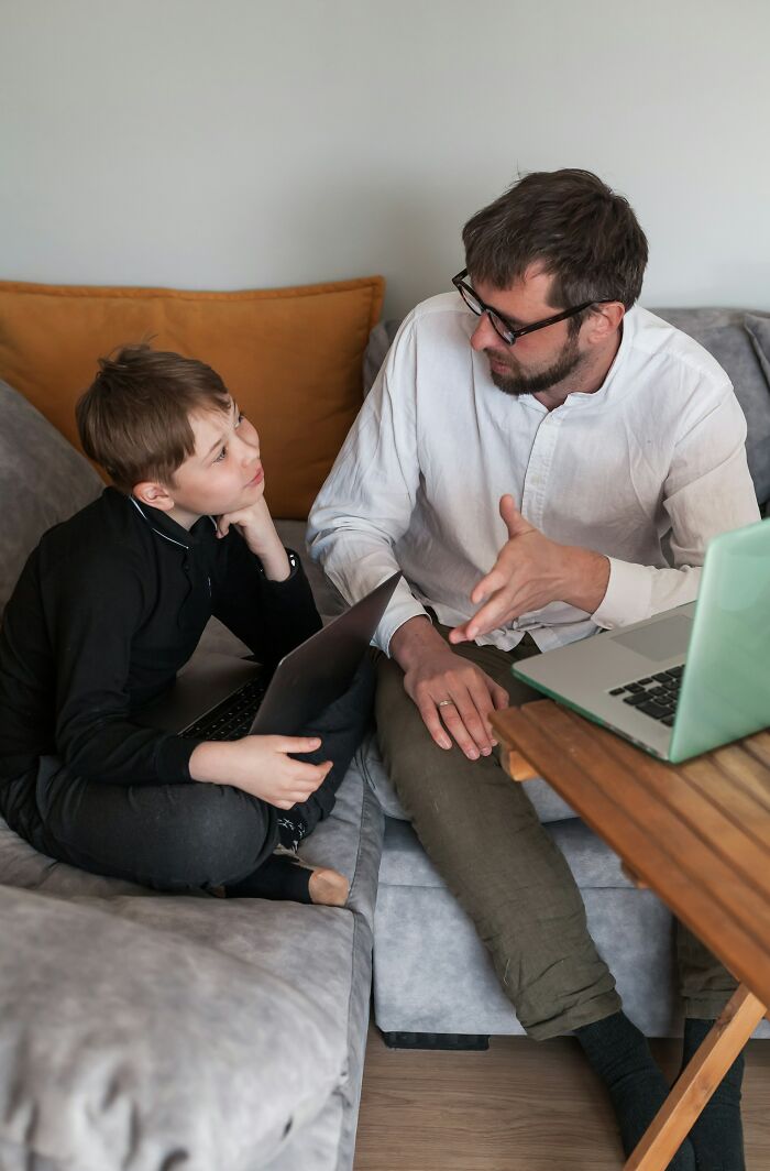 Father and son having a serious conversation on a couch with laptops, illustrating modern parenting trends in a home setting.
