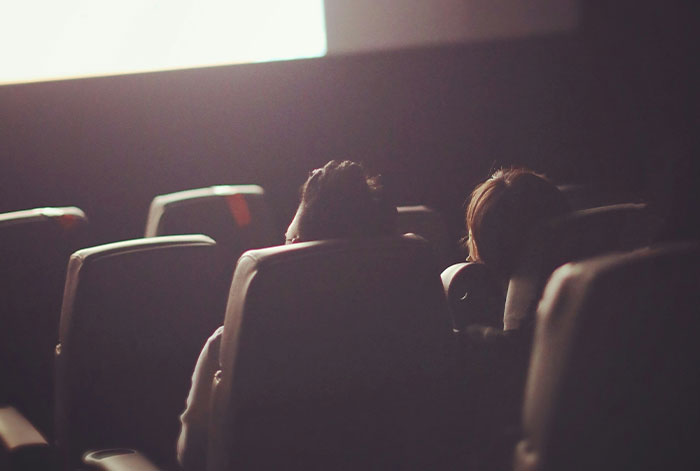Two people seated in a dimly lit theater, backlit by a large screen, conveying a tense and dramatic atmosphere.