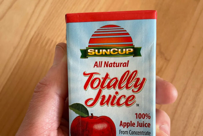 Hand holding a small carton of SUNCUP All Natural Totally Juice 100% apple juice on a wooden surface background.