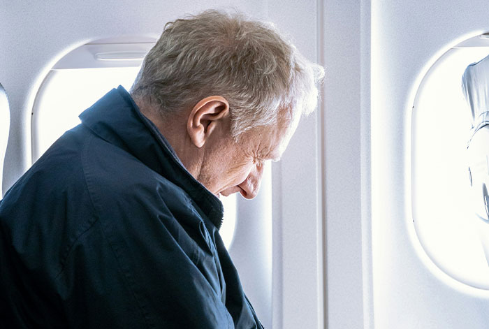 Older man in navy jacket leaning forward and closing his eyes on an airplane, illustrating lung collapse during flight emergencies.