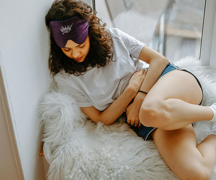 Young woman with a sleep mask holding her abdomen on a cozy chair illustrating common health concerns.