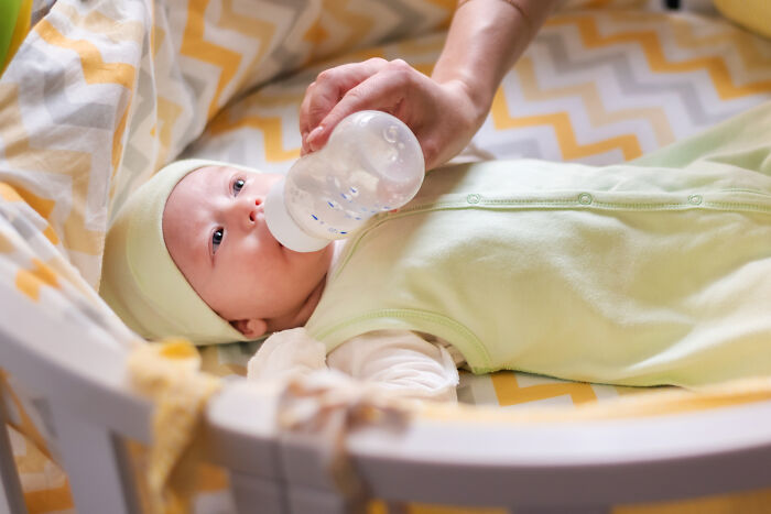 Baby lying in crib wearing green clothes, being fed from a bottle, illustrating common health concerns that freak people out but are normal.
