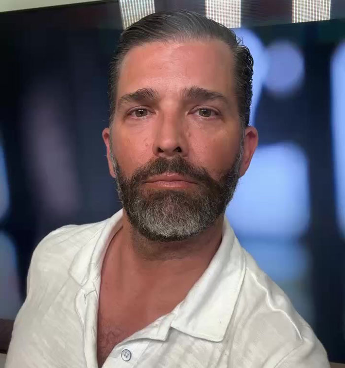 Donald Trump Jr. with a trimmed beard and white shirt, posing for a close-up photo indoors.