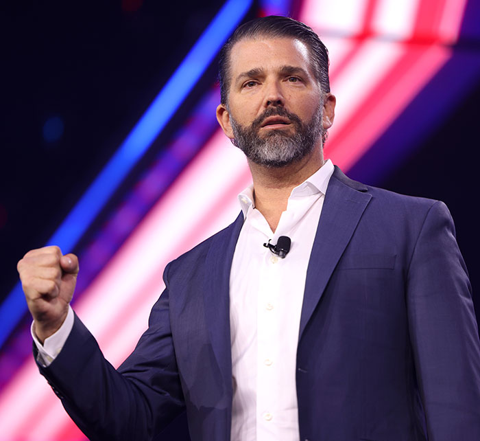 Donald Trump Jr. speaking at an event, wearing a navy suit and white shirt with a microphone on stage.