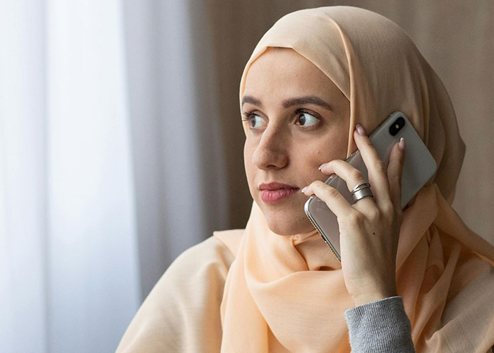 Young Muslim woman wearing a hijab, holding a smartphone, looking concerned while having a phone conversation at home.
