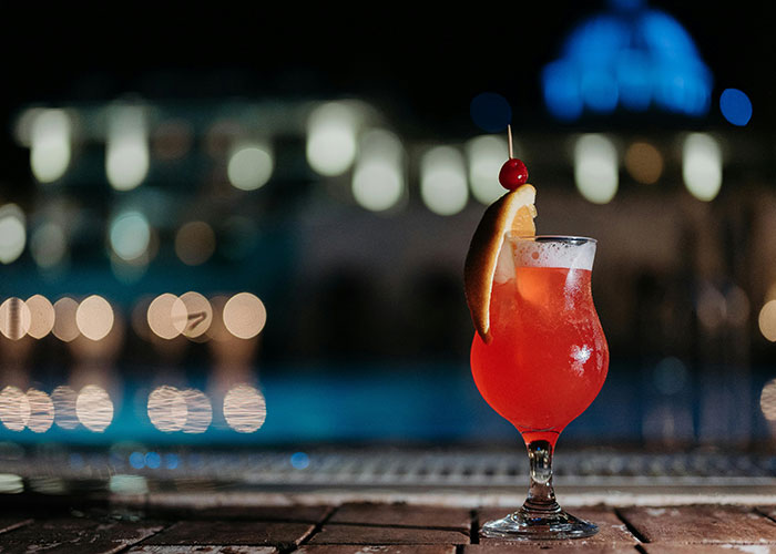 Red cocktail glass with fruit garnish by pool at night, illustrating family conflict over Muslim DIL dress code at bridal party.