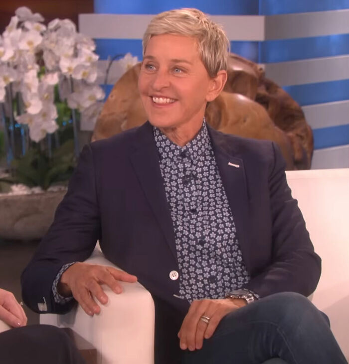Comedian on a talk show set, smiling and wearing a patterned shirt and blazer, discussing banned stars from Tonight Show.