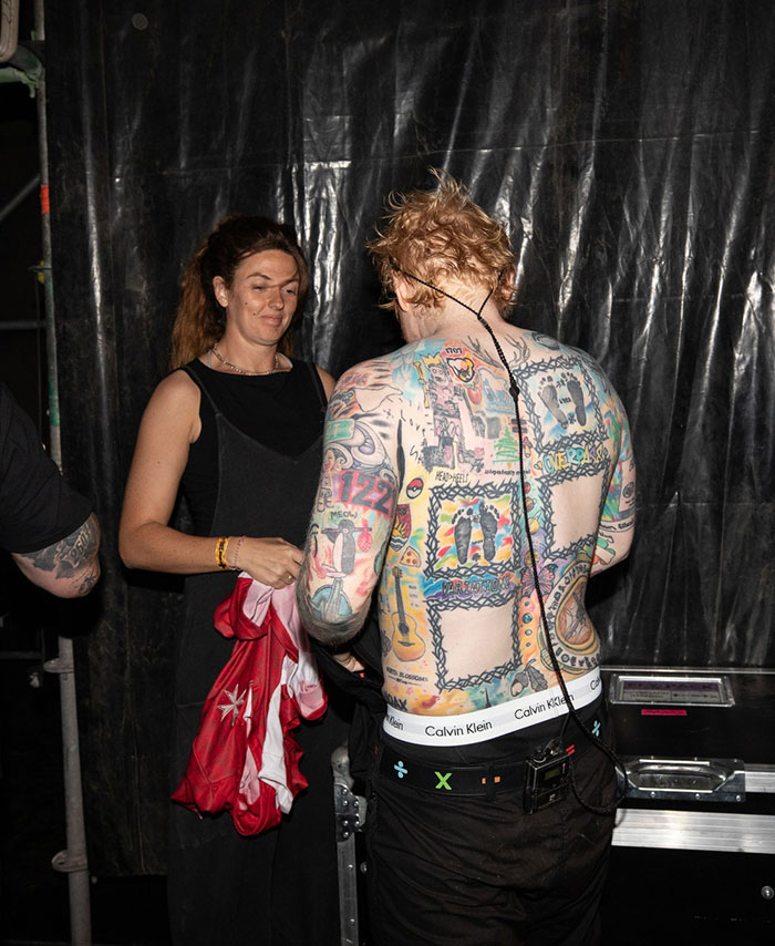 Ed Sheeran showing his shredded physique and full back tattoos backstage wearing black pants and white Calvin Klein underwear.
