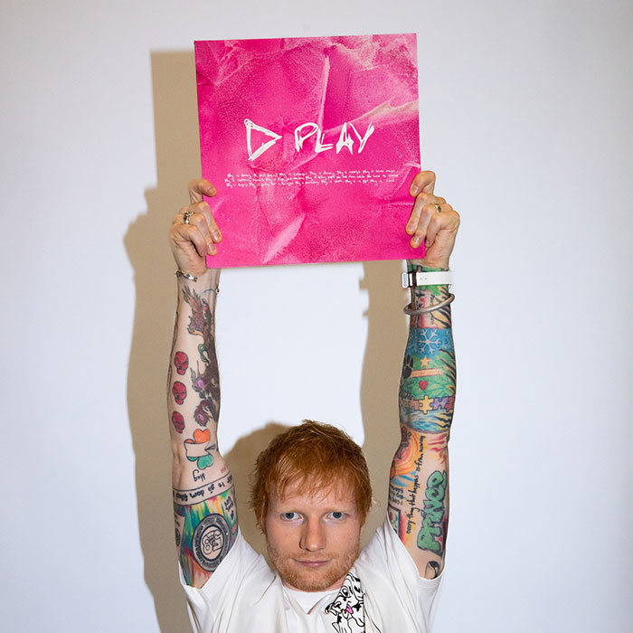 Ed Sheeran showing tattoos on both arms, holding a pink album cover above his head against a plain background