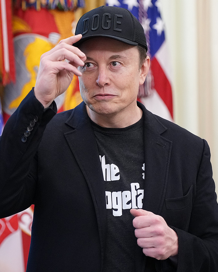 Elon Musk wearing a black Doge hat and jacket, making a gesture with his hand in front of flags indoors.