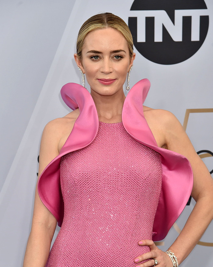 Emily Blunt in a pink textured dress with ruffled shoulders, attending an event amid plastic surgery rumors and surgeon debate.