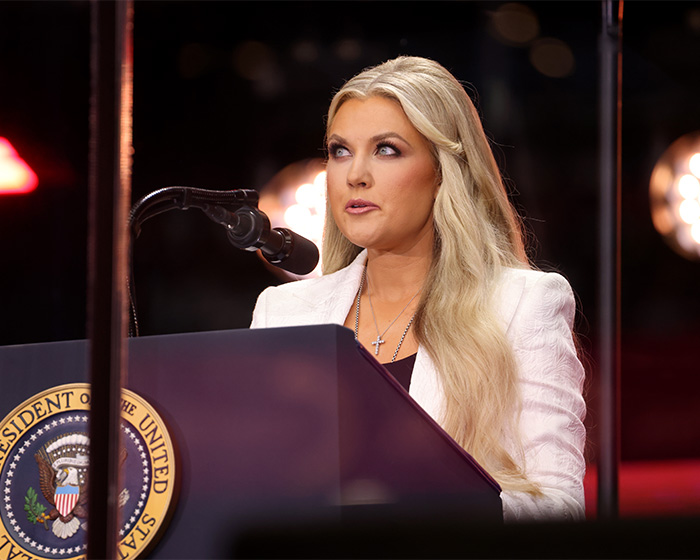 Erika Kirk speaking at a podium with the presidential seal, accused of cashing in on tragedy with Christmas message.