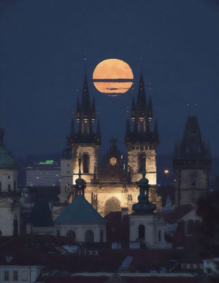 Gothic buildings with pointed spires illuminated at night under a large orange moon, evoking evil horror movie architecture.