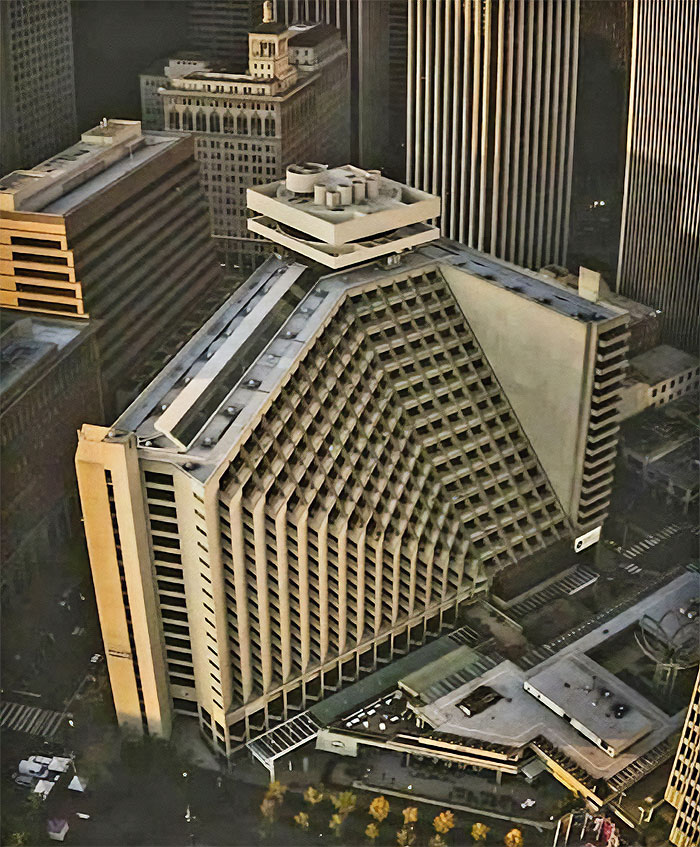 Aerial view of an evil building with a triangular, brutalist design in a dense urban cityscape at dusk.