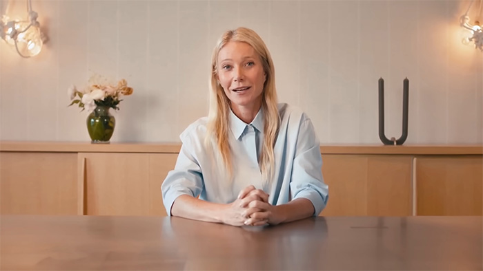 Gwyneth Paltrow seated at a table in a light blue shirt, speaking in a minimalist room with flowers and modern decor.