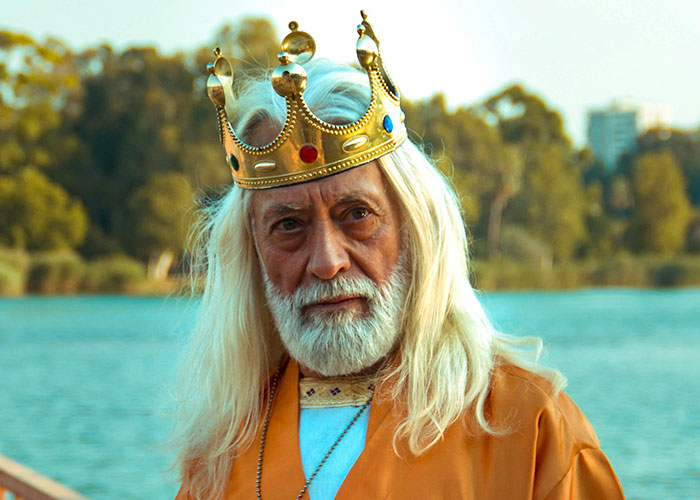 Older man with long white hair and beard wearing a golden crown by a lake, illustrating moments people share in a cult.
