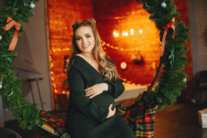 Young new mom in a cozy holiday setting, smiling while sitting inside a large Christmas wreath swing.