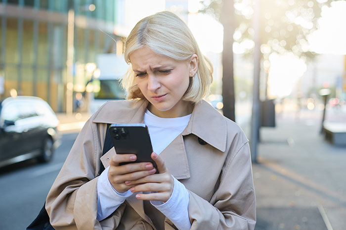 Woman looking upset while checking her phone outdoors, reflecting mom furious after dad and stepmom plan Disney trip excluding her.