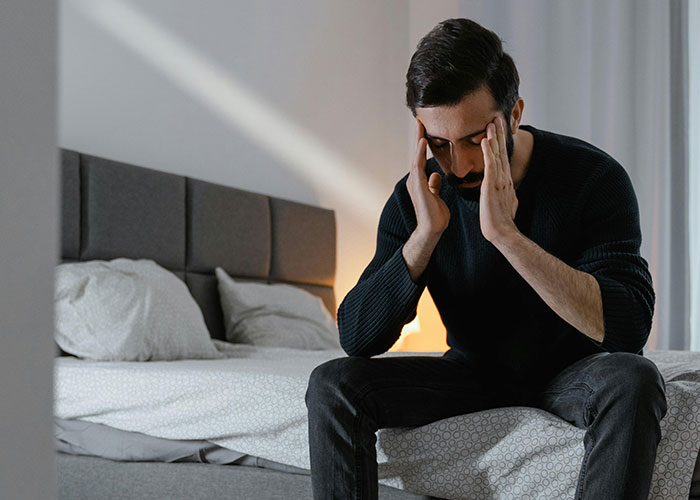 Man sitting on edge of bed holding his head, appearing stressed and overwhelmed by fianc&eacute;e&rsquo;s unexpected wedding request.