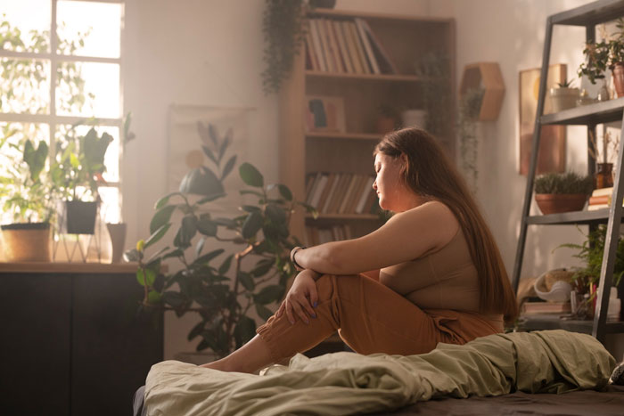 Teen girl sitting on bed in a sunlit room, reflecting quietly surrounded by plants and books, highlighting health and family care. Teen girl sitting on bed in a sunlit room, reflecting quietly surrounded by plants and books, highlighting health and family care.
