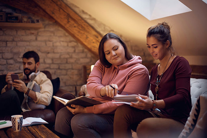 Two friends sitting together indoors, one girl playfully roasting another while the other listens and smiles.