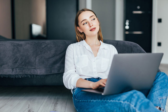 Young woman sitting on floor with laptop looking away, reflecting on host telling friend&rsquo;s wife about inviting random people.