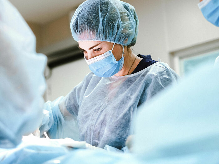 Medical staff in surgical gowns and masks focused on a procedure with patients coming off anesthesia in the background.