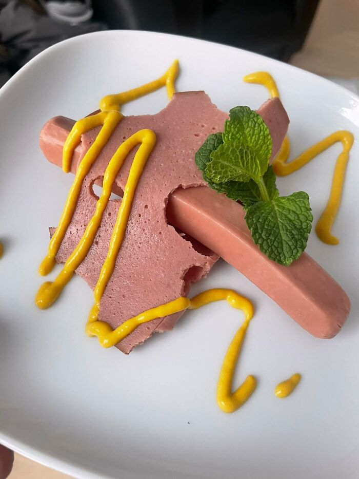 Close-up of a poorly presented food dish with processed meat, mustard squiggles, and a sprig of mint on a white plate.