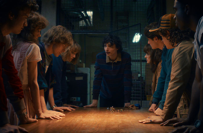 Group of Stranger Things Season 5 characters leaning over a table, discussing intense plans in a dimly lit room.