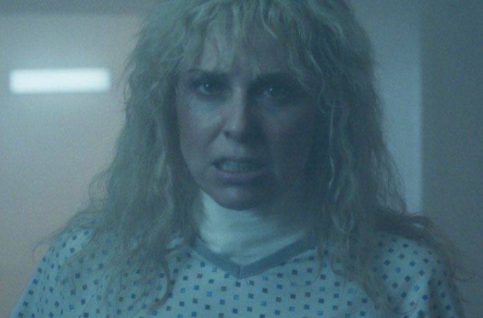 Woman with curly blonde hair wearing a hospital gown, appearing intense in a scene from Stranger Things Season 5.