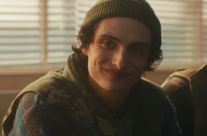 A young man wearing a beanie and camo jacket smiling during a Stranger Things Season 5 scene.