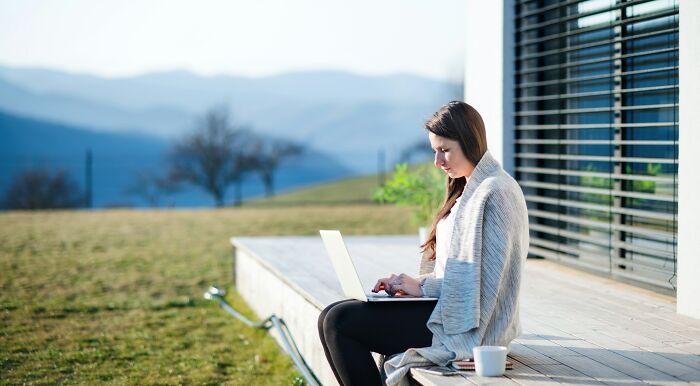 Woman working on laptop outdoors wrapped in a blanket, showing the digital nomad lifestyle with scenic mountain views.