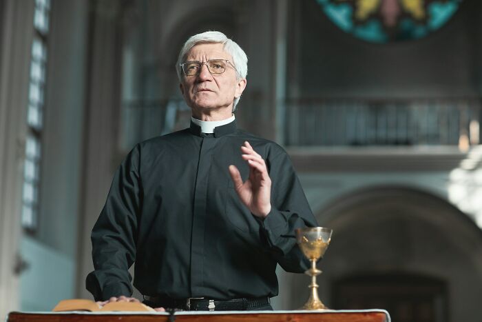 Priest in black clerical attire conducting a funeral ceremony inside a dimly lit church setting.